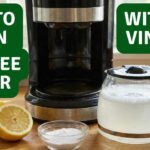A black drip coffee maker on a wooden kitchen counter next to fresh cut lemons, a small bowl of baking soda, and a glass carafe filled with a bubbly cleaning solution.