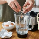 Close-up of hands crushing eggshells into a glass blender pitcher with water to demonstrate how to sharpen blender blades at home using the eggshell hack.