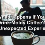 A close-up of a white coffee mug filled with dark liquid topped with green mold spots, placed on a wooden table in a dimly lit cafe setting with a blurred person in the background near a window showing a "Youngstown" sign.