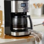 A silver drip coffee maker leaking brown water from the bottom onto a kitchen counter, with a hand holding a screwdriver and a towel ready to fix the leak.