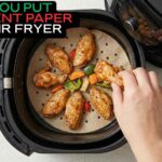 A hand placing seasoned chicken wings and vegetables on perforated parchment paper inside a black air fryer basket.