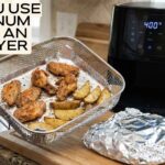 Person holding an air fryer basket lined with aluminum foil containing cooked chicken wings and potato wedges in a modern kitchen.