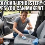 A woman inside a car using a spray bottle and scrub brush to apply one of the 5 Best DIY Car Upholstery Cleaner Recipes You Can Make at Home to a stained gray seat.