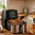 A black and silver air fryer on a wooden kitchen counter featuring a wire rack, a glass oil sprayer, a red silicone liner, and silicone-tipped tongs.