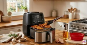 A black and silver air fryer on a wooden kitchen counter featuring a wire rack, a glass oil sprayer, a red silicone liner, and silicone-tipped tongs.
