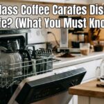 A glass coffee carafe sitting inside an open dishwasher rack and another carafe on a wooden kitchen counter with text overlay: Are Glass Coffee Carafes Dishwasher Safe? (What You Must Know).