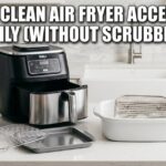 A Ninja air fryer sits on a clean white counter. It has dirty metal racks and a basket next to it. One rack is soaking in a bowl of soapy water. Blue soap and baking soda are on the table. The text says How to Clean Air Fryer Accessories Easily (Without Scrubbing).