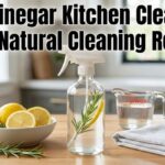 A glass spray bottle of DIY Vinegar Kitchen Cleaner: Safe Natural Cleaning Recipe on a wooden kitchen counter with fresh lemons, rosemary, and a bowl of baking soda.