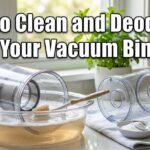 A transparent plastic vacuum bin being washed in a bowl of soapy water next to baking soda and lemon essential oil on a clean kitchen counter.