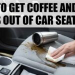 A hand blotting a fresh coffee spill on a gray cloth car seat using white paper towels next to a tipped-over silver travel mug.