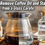 Before and after view of a glass coffee carafe showing heavy coffee oil stains on one side and a clean, clear surface on the other, with baking soda, vinegar, salt, and ice nearby.