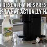 A black Nespresso machine on a kitchen counter next to a large jug of white vinegar and a measuring cup, with the text "Can You Descale a Nespresso with Vinegar? (What Actually Happens)" overlayed in Impact font.