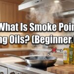 A smoking frying pan on a kitchen stove next to bottles of extra virgin olive oil, avocado oil, canola oil, and ghee, with the text "What Is Smoke Point in Cooking Oils? (Beginner Guide)" across the center.