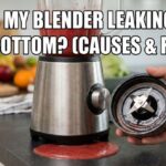 A silver blender leaking red smoothie from the bottom onto a kitchen counter, with a hand holding the removed blade assembly to inspect the rubber gasket.