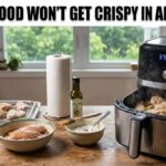 A hot black air fryer full of steaming wet food on a wood counter. Raw chicken, a fresh potato, clean paper towels, and pure oil sit near it. Bold text at the top says Why Wet Food Won’t Get Crispy in an Air Fryer.