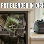 A dirty blender pitcher with green smoothie residue sitting on the top rack of an open dishwasher next to plates and mugs. The motor base sits on a white kitchen counter near a batter spill, with bold white text at the top asking, "CAN YOU PUT BLENDER IN DISHWASHER."