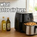 A black air fryer with an open basket full of crispy french fries on a kitchen counter next to bottles of avocado oil, light olive oil, grapeseed oil, and an oil sprayer, with the text what oil is best for air fryer.