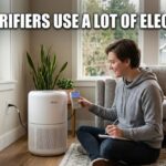 A person sitting on a living room floor using a digital wall meter to test the power draw of a white cylindrical air purifier.