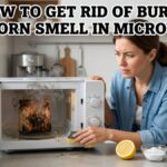 A woman in a bright kitchen looks at a smoking microwave with a burnt popcorn bag inside, with bold text "HOW TO GET RID OF BURNT POPCORN SMELL IN MICROWAVE" at the top and natural cleaning supplies like lemon and vinegar in the foreground.
