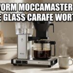 A silver coffee machine sits on a white counter. It has a glass pot with dark coffee inside. Large white text asks if the glass carafe is worth it.