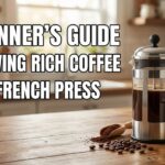A stainless steel and glass French press filled with dark coffee sits on a wooden table next to a white mug and scattered coffee beans. Large white text reads: A BEGINNER'S GUIDE TO BREWING RICH COFFEE WITH A FRENCH PRESS.