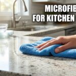 A hand wipes a shiny granite kitchen countertop with a blue microfiber cloth. A blue non-scratch sponge and glass bowls are in the background. Text overlay reads: MICROFIBER CLOTH FOR KITCHEN CLEANING.
