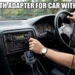 A man's hand reaching for an older car's radio dial while a Bluetooth adapter for car without aux is plugged into the 12V cigarette lighter, with a smartphone in a dashboard mount displaying a music app.