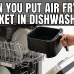 A person placing a black, square air fryer basket into the bottom rack of an open dishwasher with large white text asking "Can you put air fryer basket in dishwasher?".