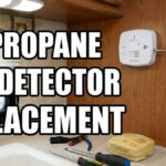 A person using a screwdriver to install a white RV propane detector on a wooden wall above a camper kitchen sink, featuring large bold text reading RV PROPANE GAS DETECTOR REPLACEMENT.