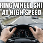 A first-person view of a driver's hands on a steering wheel while traveling fast on a highway. The image features motion blur and large white text that reads "STEERING WHEEL SHIMMY AT HIGH SPEED".