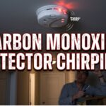 A frustrated man in pajamas, holding a screwdriver on a stepladder, looks up at a ceiling-mounted carbon monoxide detector which has flashing red lights and sound wave lines indicating a chirp.