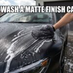 A close-up shot of a hand using a grey microfiber wash mitt to clean the hood of a matte black car covered in white soapy suds, with two wash buckets visible in the driveway and bold text overlaying the top.