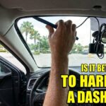 A man inside a car sweating while prying at the windshield trim to hide dash cam wires and holding a flashlight in his mouth.