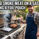 A smiling American man in a blue apron standing next to an open gas grill on a backyard deck, with smoke rising from a foil packet and meat cooking on the grates.