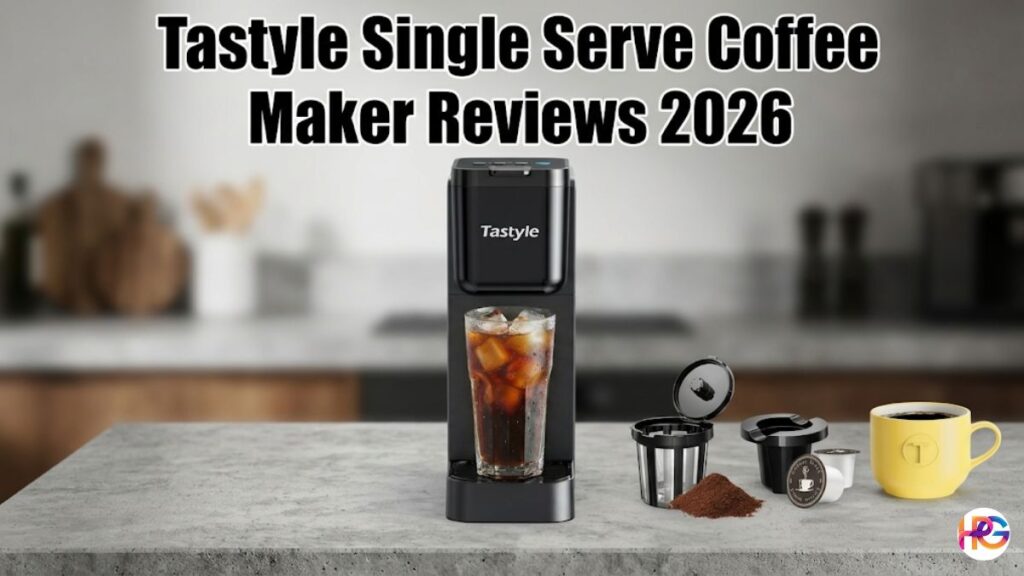 A wide banner graphic titled "Tastyle Single Serve Coffee Maker Reviews 2026" featuring a slim black Tastyle coffee brewer sitting on a gray kitchen countertop. A tall glass of iced coffee is under the spout, while a reusable filter basket, ground coffee, a coffee pod, and a yellow mug sit to the right side against a blurred kitchen background.