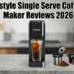 A wide banner graphic titled "Tastyle Single Serve Coffee Maker Reviews 2026" featuring a slim black Tastyle coffee brewer sitting on a gray kitchen countertop. A tall glass of iced coffee is under the spout, while a reusable filter basket, ground coffee, a coffee pod, and a yellow mug sit to the right side against a blurred kitchen background.