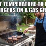 A smiling Caucasian man in a blue polo shirt and black apron cooks burgers on a modern Weber gas grill in a backyard. Large white Impact font text across the top reads: "WHAT TEMPERATURE TO COOK BURGERS ON A GAS GRILL".
