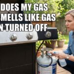 An American woman in a denim shirt kneels on a wooden patio next to a gas grill, looking concerned as she uses a soapy water spray bottle to test the propane tank valve for a leak.