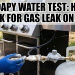A gloved hand uses a paintbrush to apply soapy water to a gas regulator and hose connected to a propane tank on an outdoor barbecue grill. Big bold text at the top reads: "THE SOAPY WATER TEST: HOW TO CHECK FOR GAS LEAK ON GRILL".