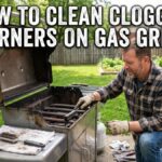 A middle-aged Caucasian man in a plaid shirt and work gloves kneels in a suburban backyard patio, using a stainless steel brush to clean the burner tubes of an open gas grill. Large text overlays the image at the top, reading: "HOW TO CLEAN CLOGGED BURNERS ON GAS GRILL". A clean towel on the side tray holds another brush and a paperclip.