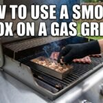 A photo shows a person wearing a black heat-resistant glove setting a small stainless steel smoker box filled with wood chips onto the flavorizer bars of a gas grill between the cooking grates, near lit burners. White text at the top of the image reads: "HOW TO USE A SMOKER BOX ON A GAS GRILL." The background is a green lawn.