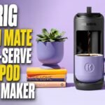 Keurig K-Mini Mate Single-Serve K-Cup Pod Coffee Maker brewing coffee into a purple mug on a countertop with decorative books, a succulent plant, and a colorful picture frame in the background.